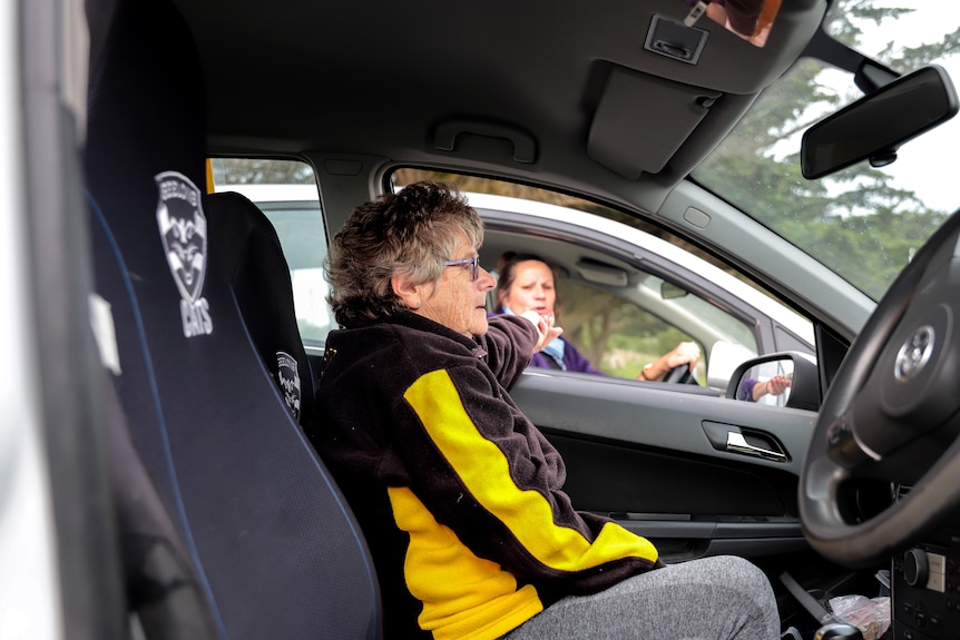 Older woman in brown and gold jumper in her car taking money from a woman in a passing car
