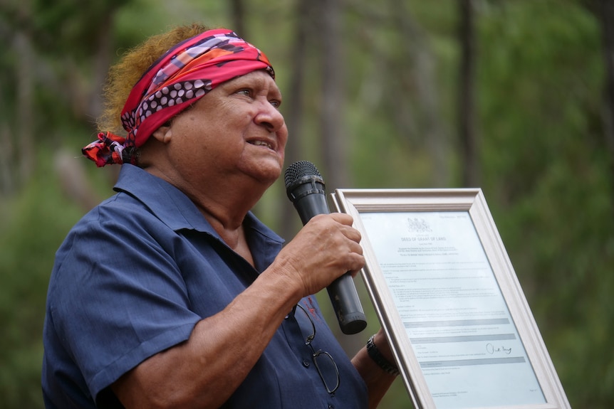 An Indigenous woman passionately speaking into a microphone, holding a deed, trees behind her.