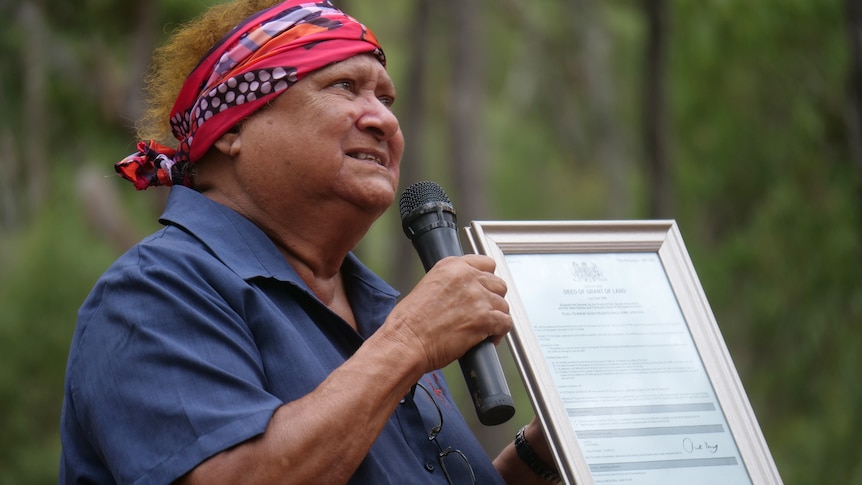 Aunty Sally passionately speaking into a microphone, holding A4 deed, trees behind her. 