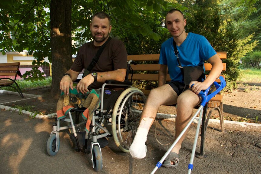 Anatoly, who is in a wheelchair, and Bogdan, who is on crutches, both smile at the camera.