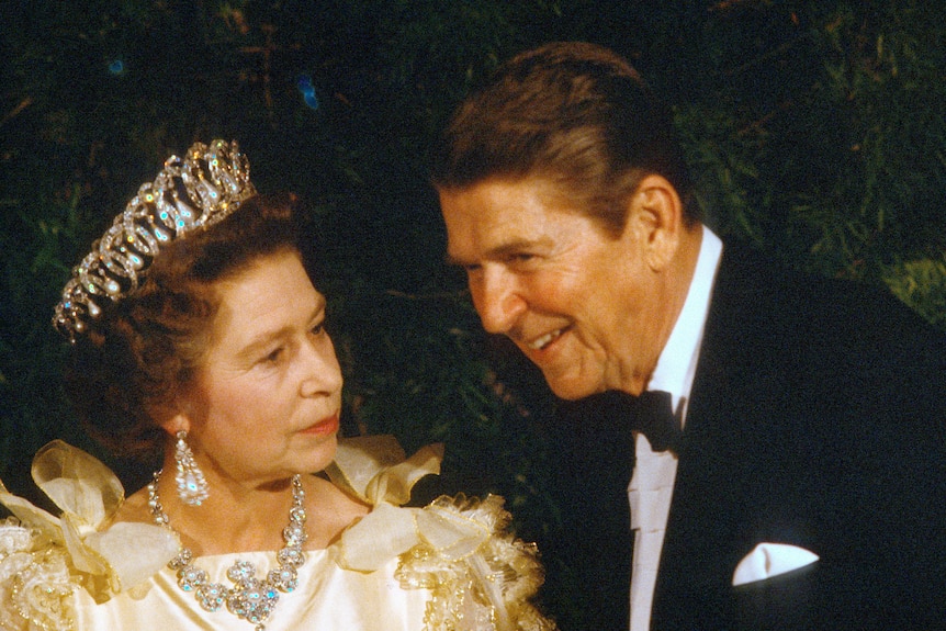 The queen in a tiara and jewells speaking with a ban with brown hair wearing a tuxedo