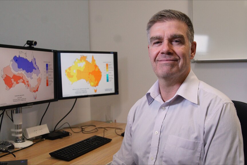 Greg Browning BOM Senior Meterologist sits at desk with weather maps behind him
