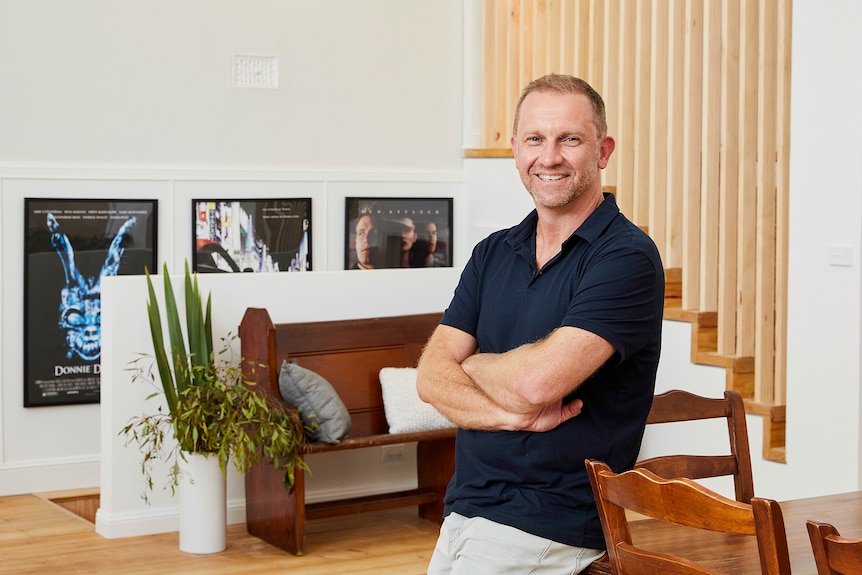 A smiling man wearing a black polo shirt leaning against a dining table in a well-lit living room