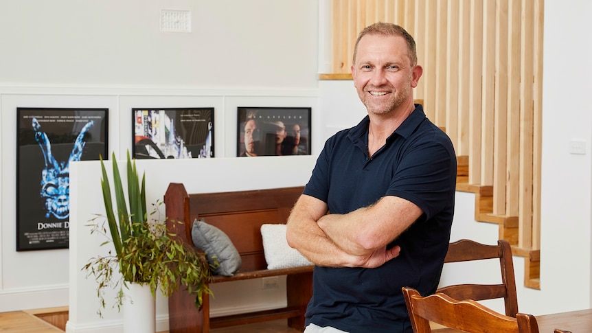 Anthony Burke wearing a black polo shirt leaning against a dining table in a well-lit living room