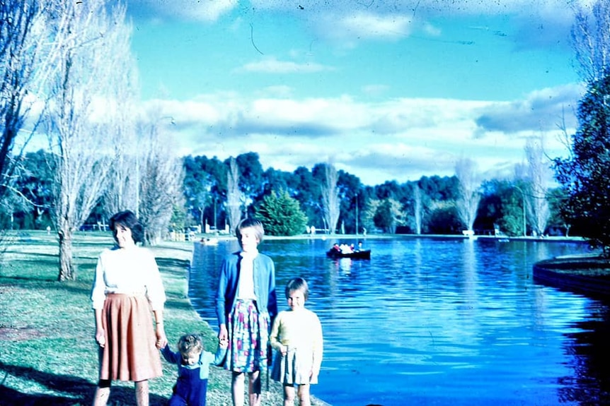 Four girls walk along grass next to a lake with a boat in the far ground.