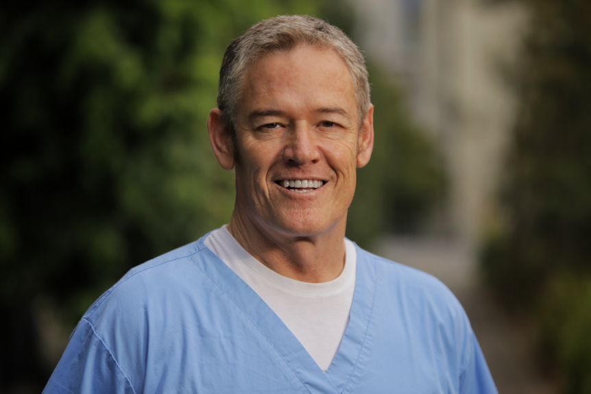 Doctor wearing light blue scrubs, standing outside. 