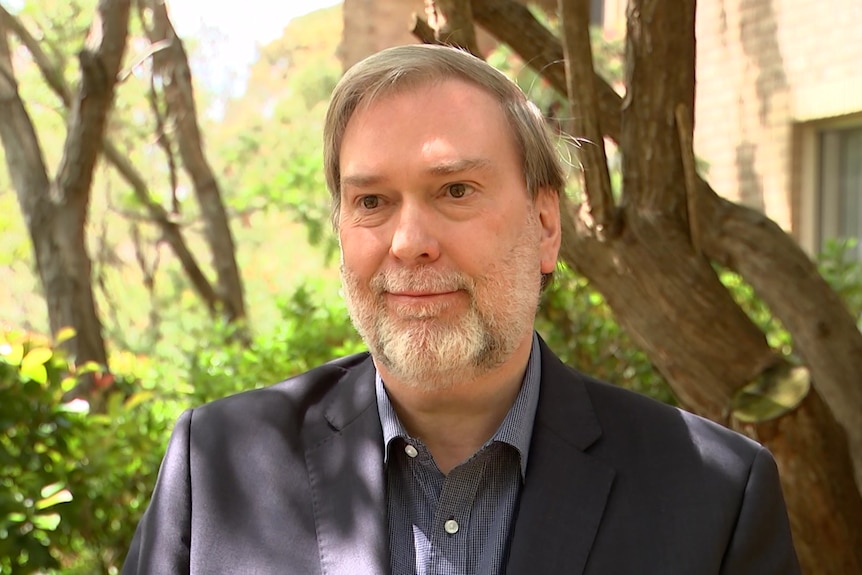 A head and shoulders shot of a grey haired, bearded man standing in front of trees