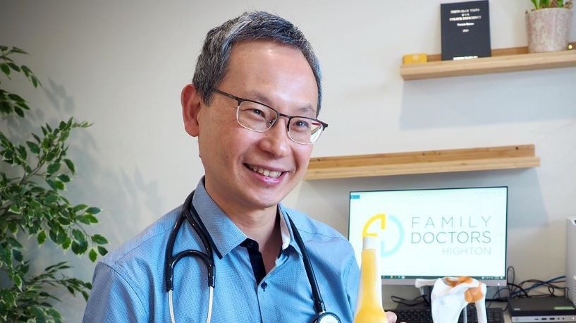 Dr Ern Chang wears a stethoscope around his neck, sits at a desk and shows a patient a model of a knee joint.