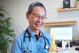 Dr Ern Chang wears a stethoscope around his neck, sits at a desk and shows a patient a model of a knee joint.