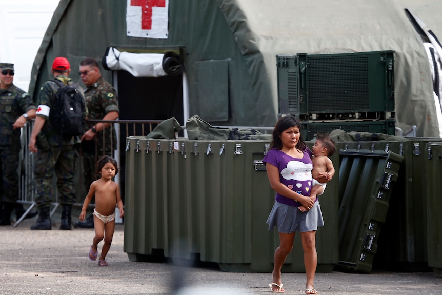 a woman carries a baby as another baby walks behind her outside an army field hospital