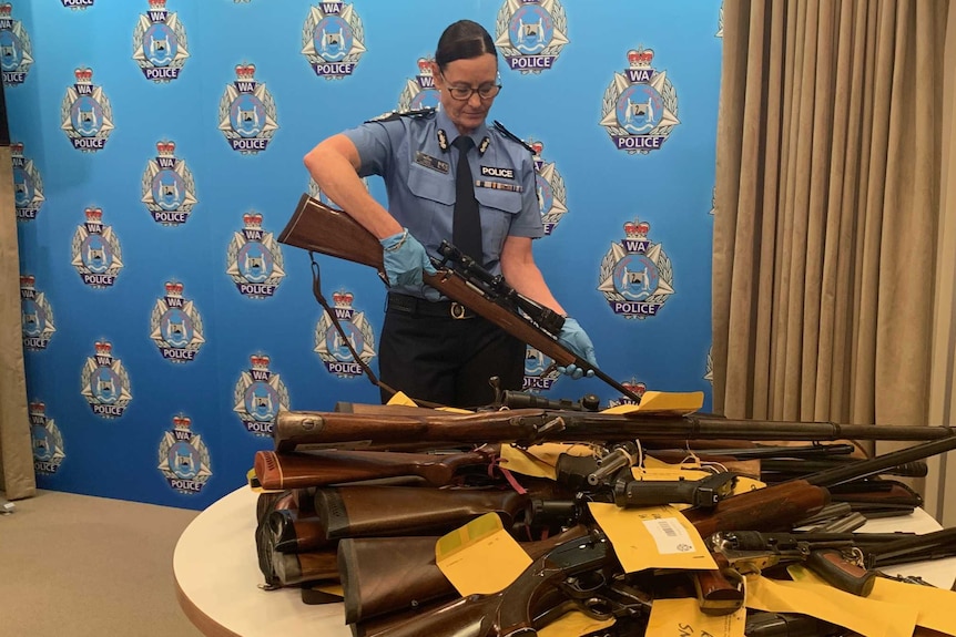 Jo McCabe holding a rifle behind table with pile of firearms on it