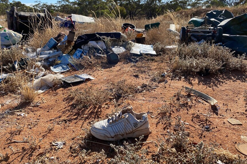 A shoe lies in brown soil amongst scrub, which is filled with rubbish behind it.