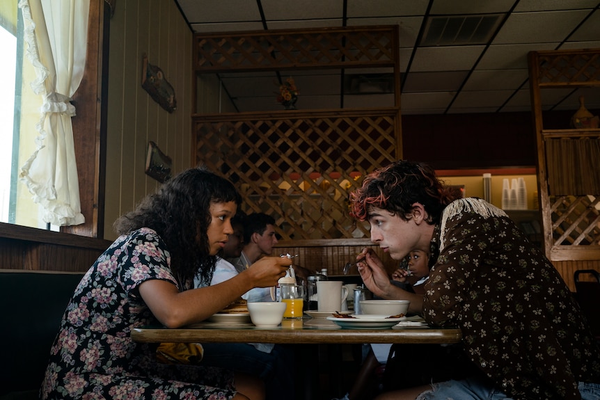 Uma jovem negra e um jovem pálido com cabelo tingido de vermelho sentam-se frente a frente em um restaurante, comendo