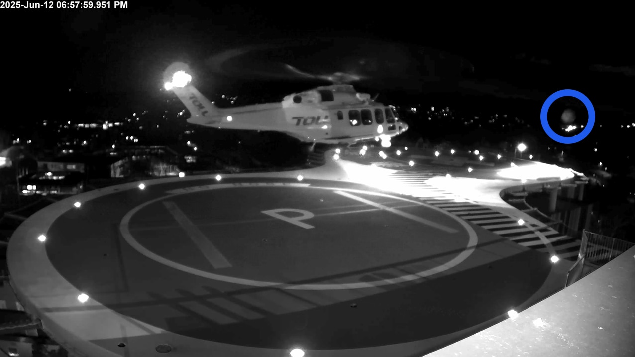 A still from black and white CCTV vision capturing an item flying through the air as a helicopter departs from its helipad.