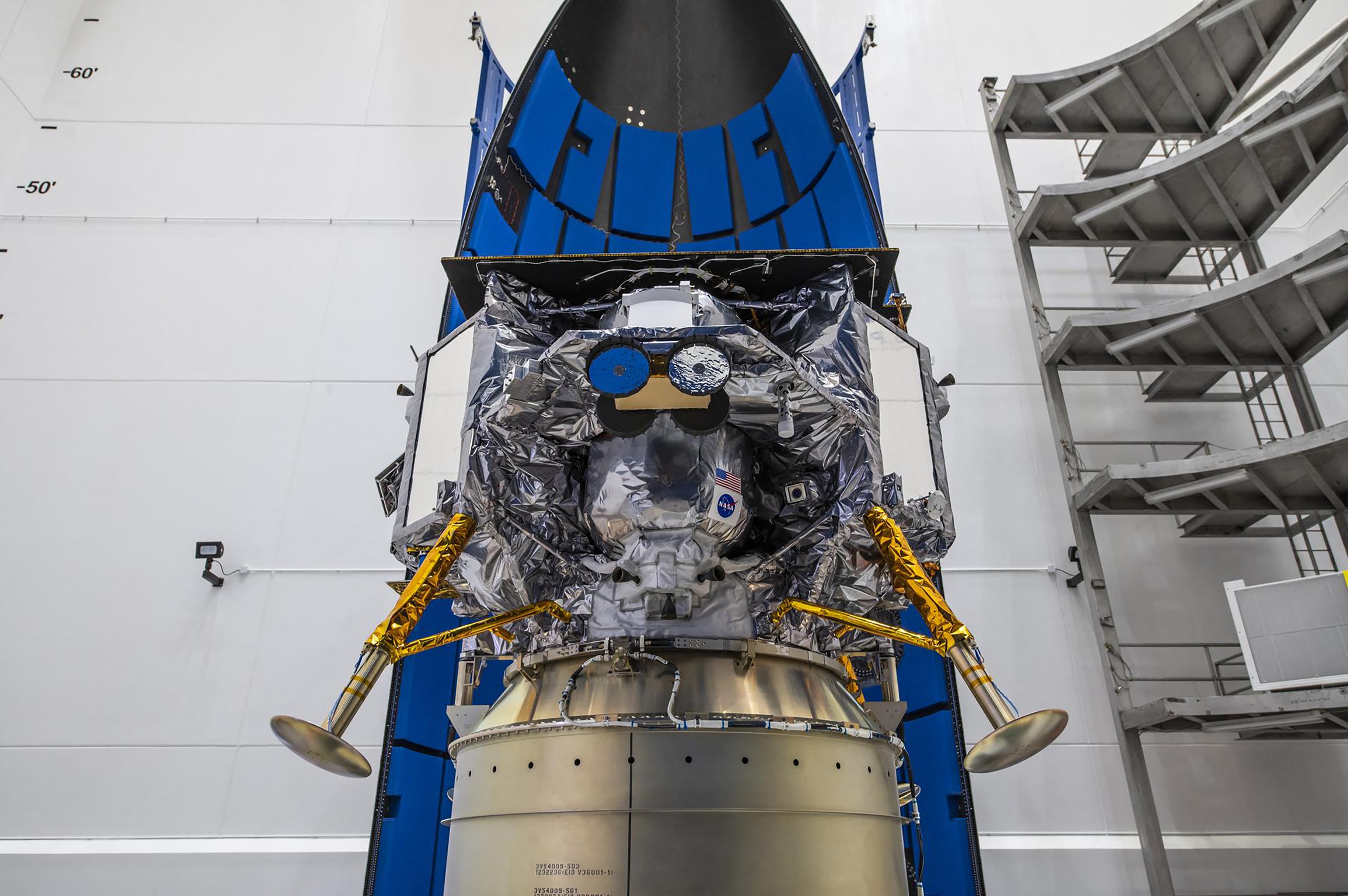 A close up of a lunar lander encapsulated in an open nose cone of a rocket, inside a factory.
