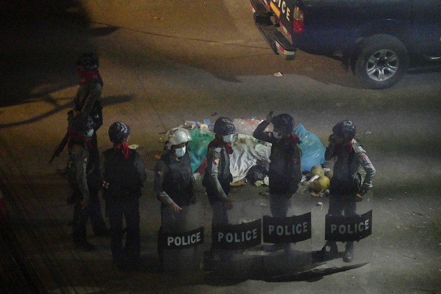 Four police officers in riot gear hold shields in front of vehicles on a poorly lit road at night.