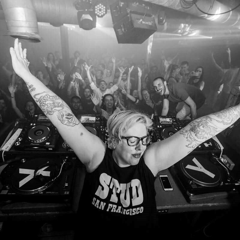 The Black Madonna, standing behind the desks at a dark club with her hands in the air, crowd also have their hands in the air