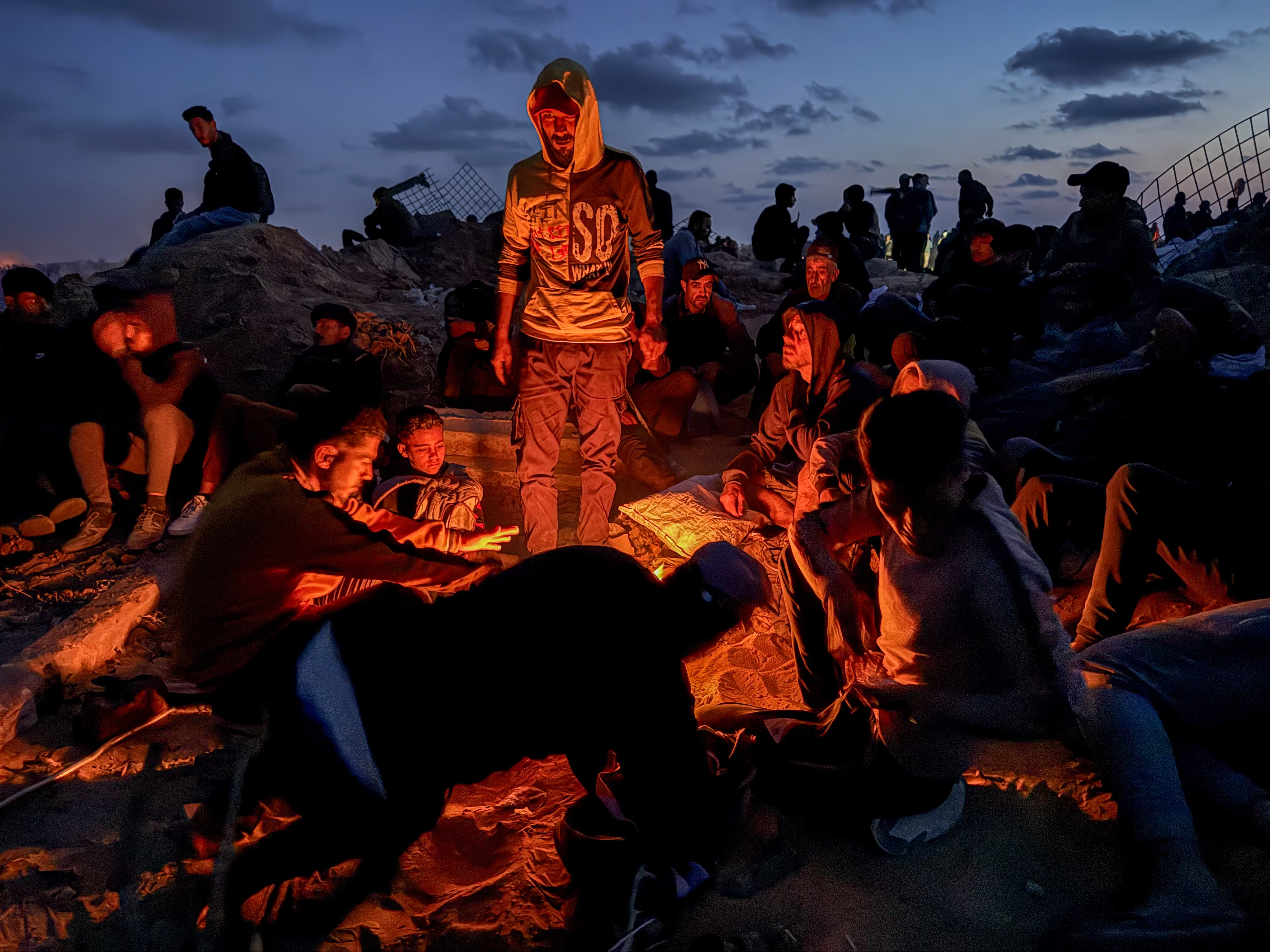 A group of men, some standing while others sit or lie on barren ground, look at the orange glow of a fire under a dusk sky.
