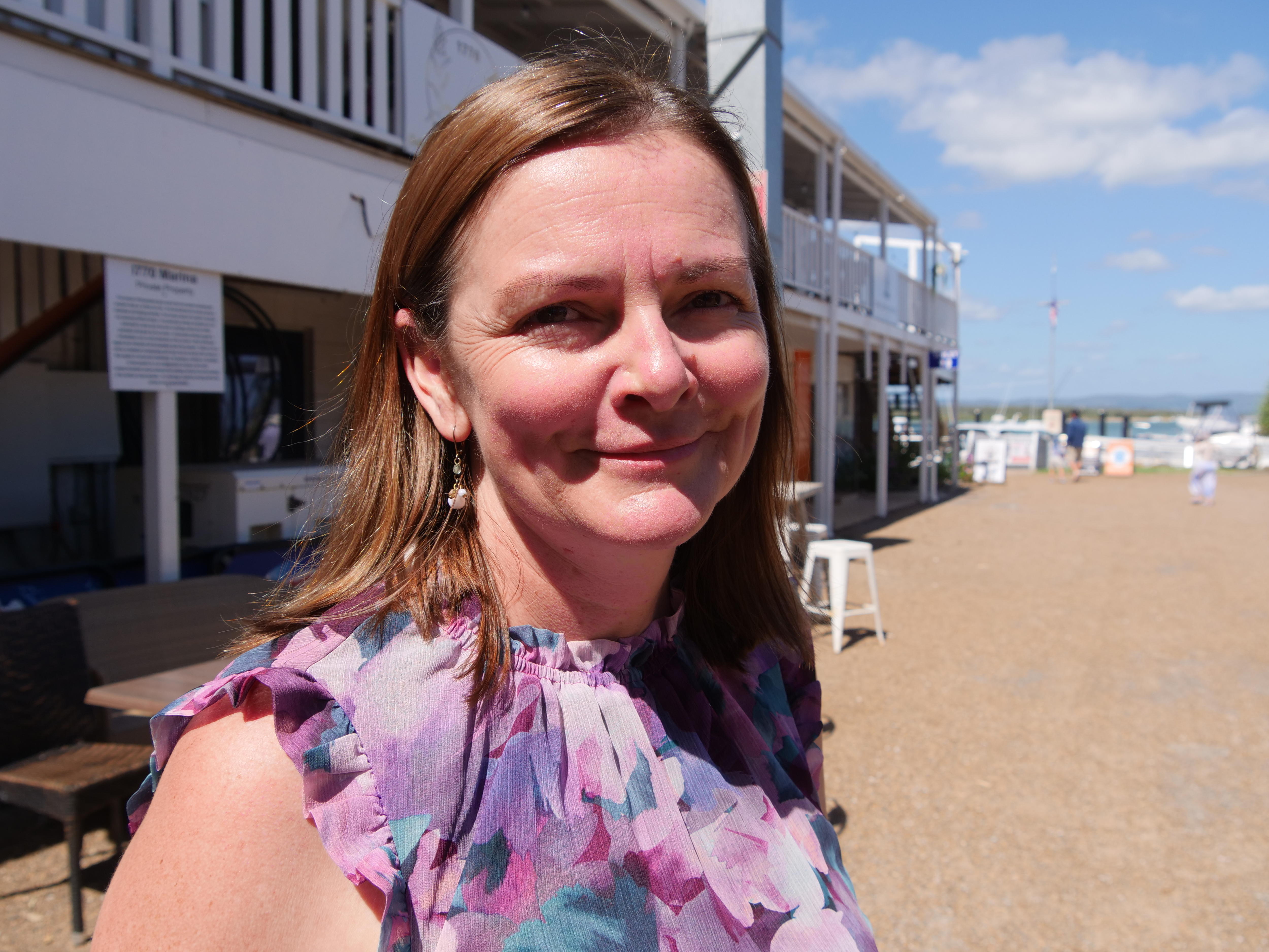 Mujer sonriendo suavemente a la cámara, vistiendo un top rosa-violeta, tiendas locales al fondo.