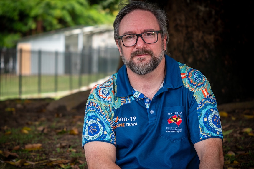 A man wearing glasses and a blue shirt decorated with Indigenous art sits under a tree.