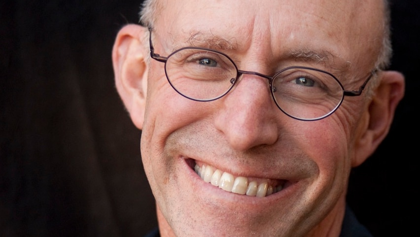From the shoulders up, a bald man wearing dark blue shirt and thin-framed glasses smiles widely.