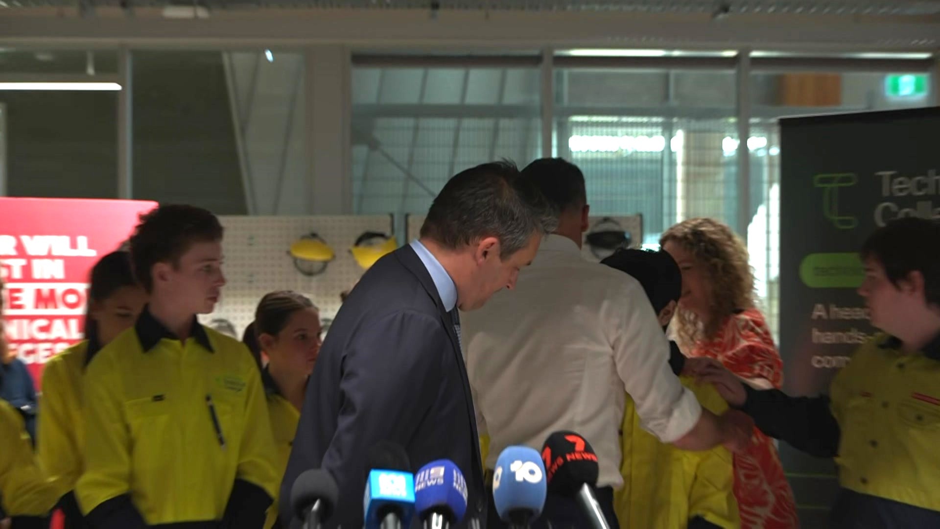 pessoas observando um adolescente apoiado em Peter Malinauskas e sendo conduzido de costas na frente dos microfones.