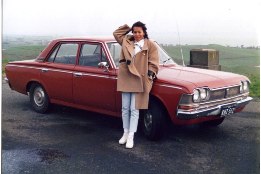 You view a young Filipina leaning against a vintage red sedan, as green rolling hills stretch out behind her to the horizon. 