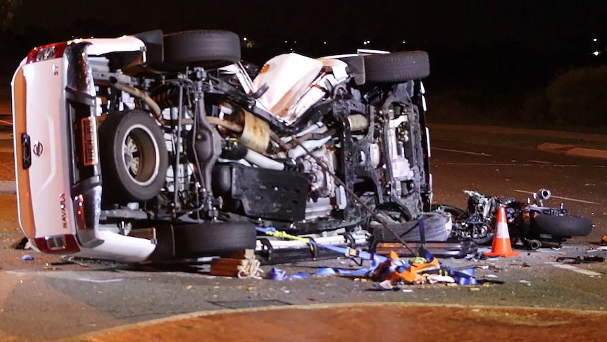A white ute lies on its side after a crash with its undercarriage visible and a wrecked motorbike lies in front of the vehicle.
