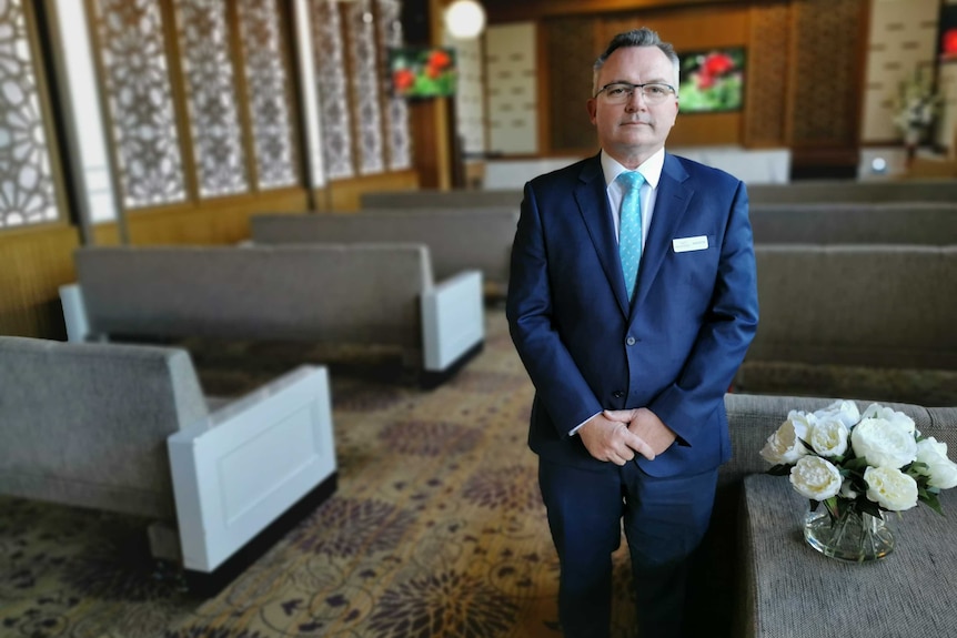 Andrew Pinder stands by a vase of white flowers, amid rows of seats.