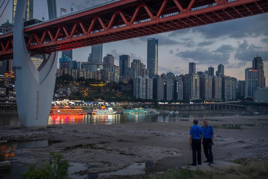 Security officers stand on a hillside after clearing away visitors from a dry riverbed, with the lights of a megacity behind.