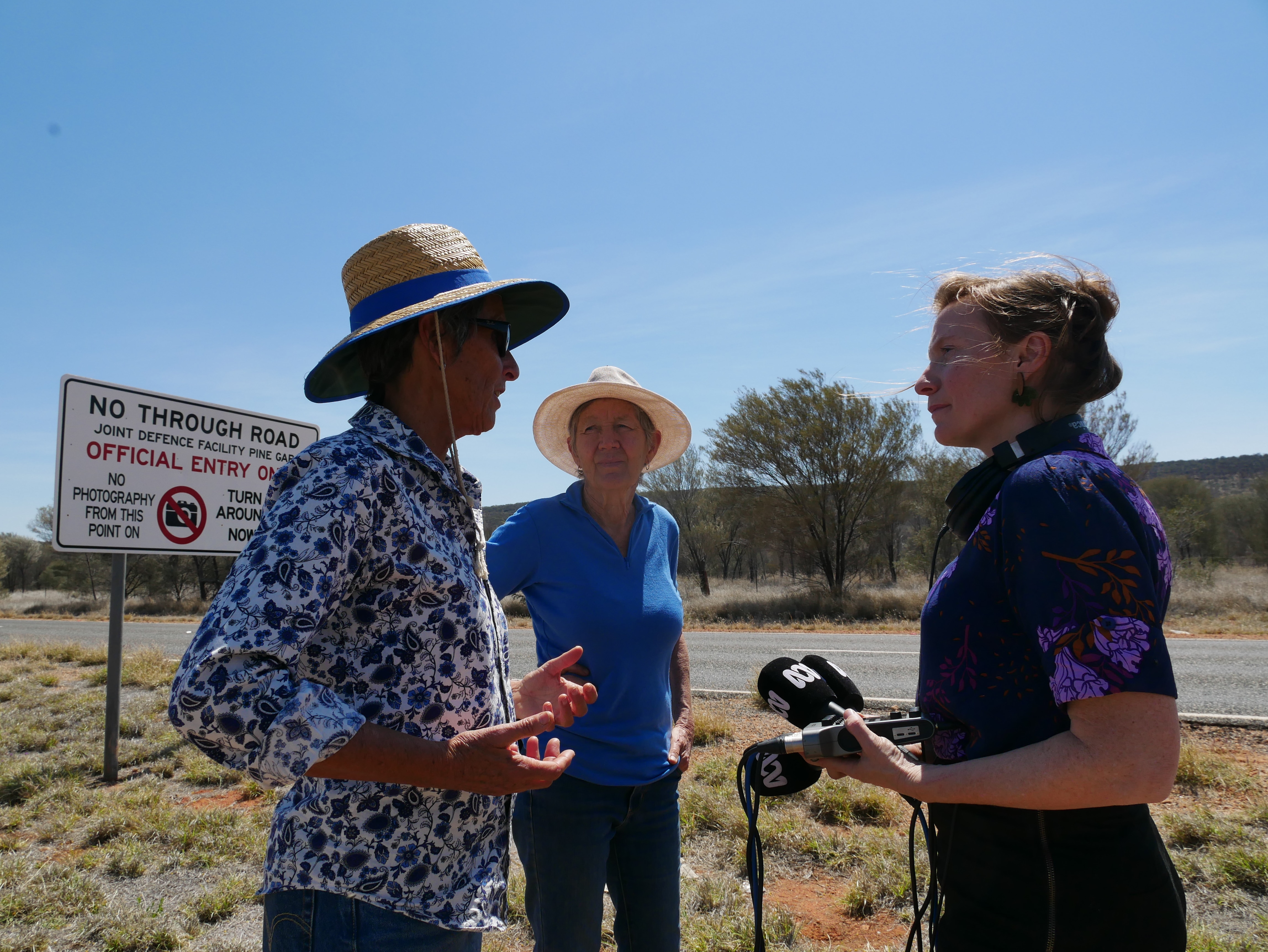 Three women stand in a huddle with one woman holding a microphone. They are outside on a sunny day.