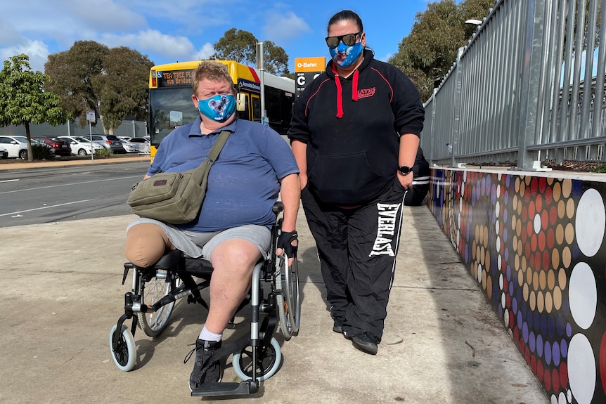 A man in a wheelchair with an amputated leg next to a woman with black hair on a sloped footpath while a bus goes behind them