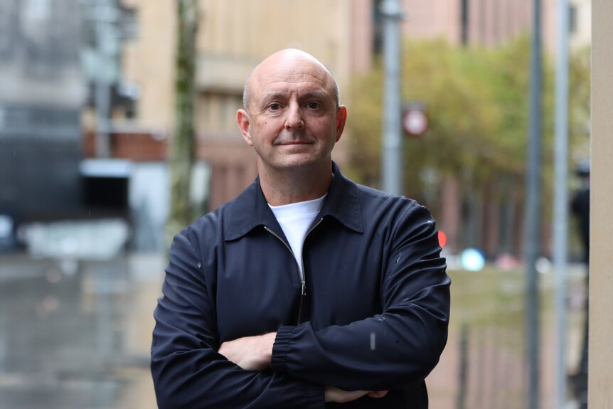 A bald white middle aged man wears a blue jacket over a white t-shirt, arms folded he stares into the camera.