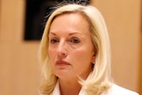 A woman with blonde hair in a formal white jacket sitting a table in front of a microphone