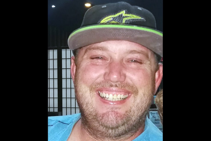An image of a smiling man with facial hair wearing a black and green cap and blue shirt.