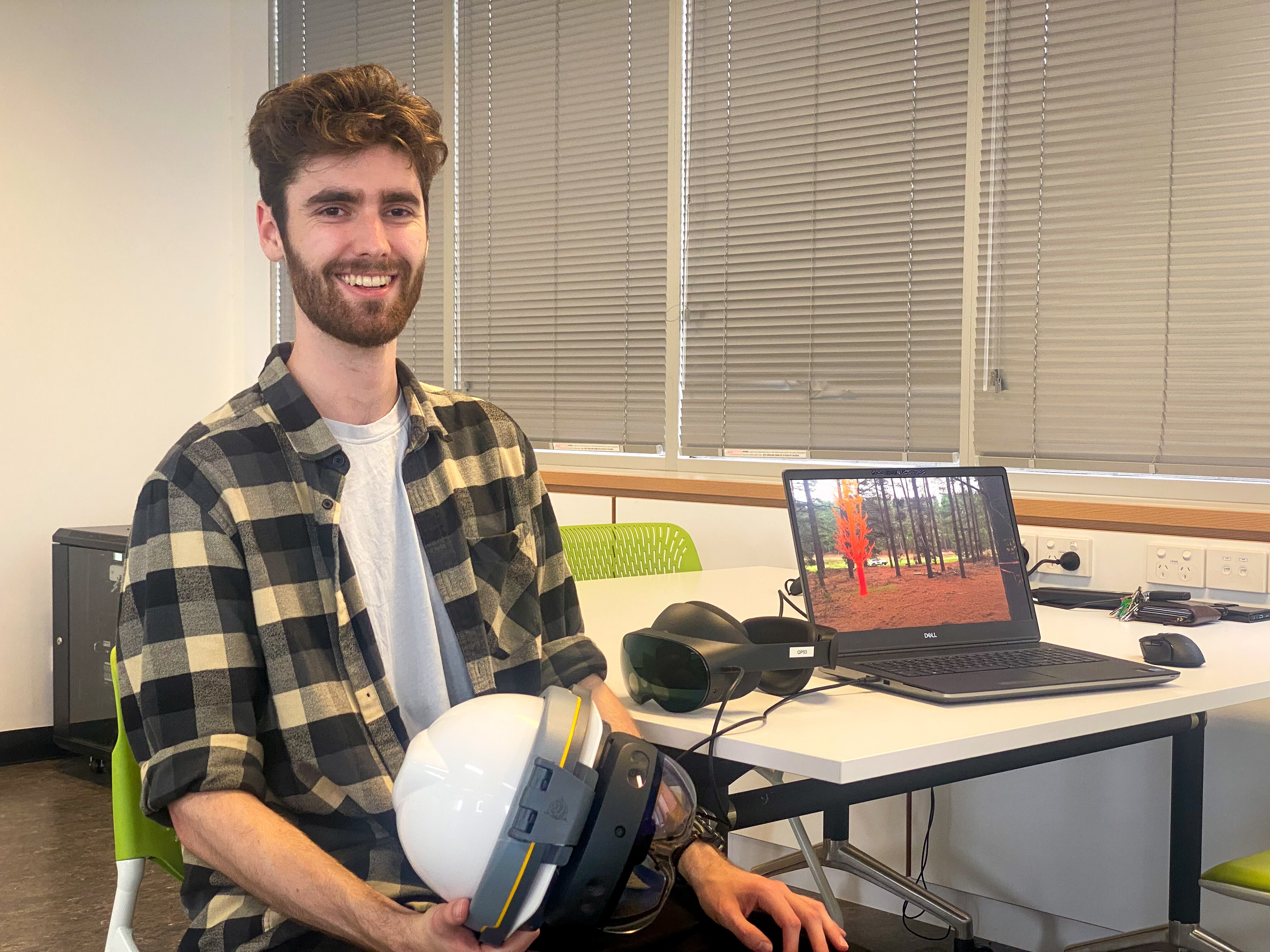 A man holds a virtual reality enabled helmet smiling at the camera, with a laptop on a desk behind him showing a virtual tree. 
