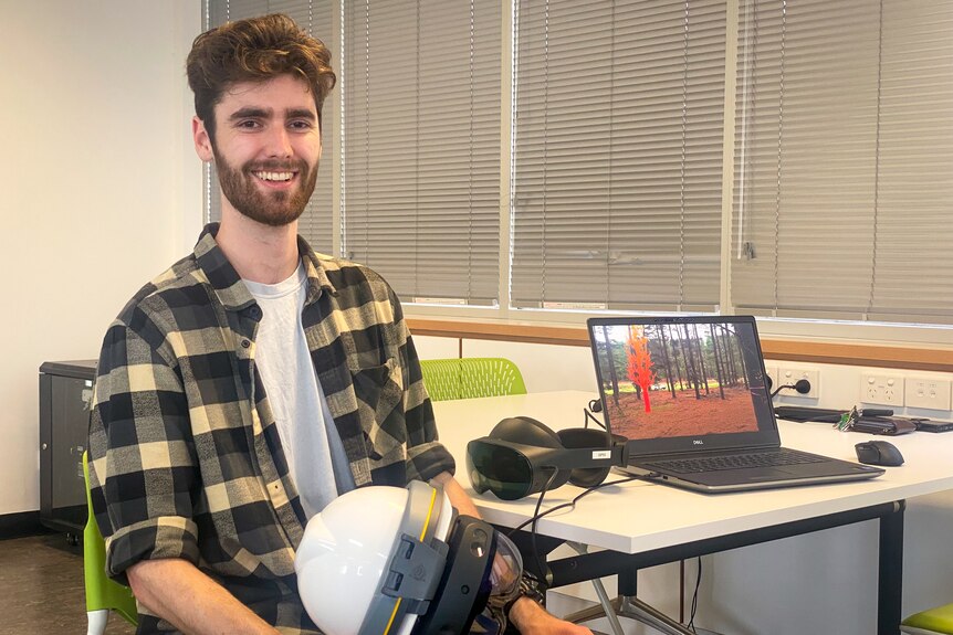 A man holds a virtual reality enabled helmet smiling at the camera, with a laptop on a desk behind him showing a virtual tree. 
