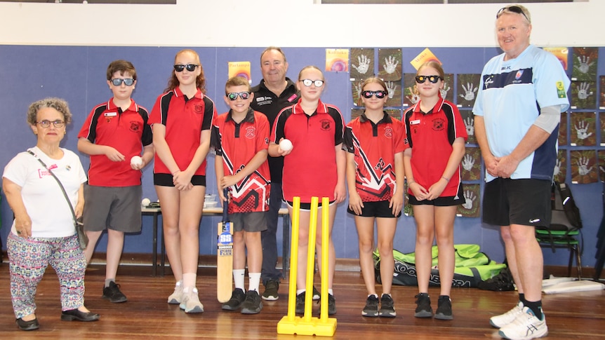 A woman with dwarfism and two men standing with a group of young students wearing glasses and holding cricket equipment.