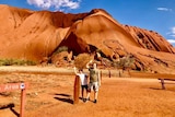 Two boys stand in front of large rock landmark under blue sky.
