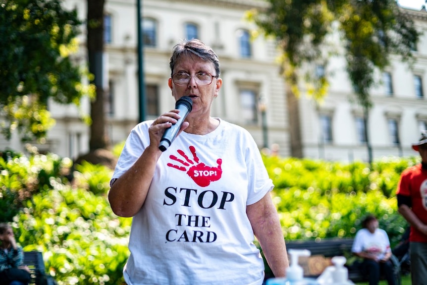 an old woman outside with a microphone in a white shirt that reads stop the card 