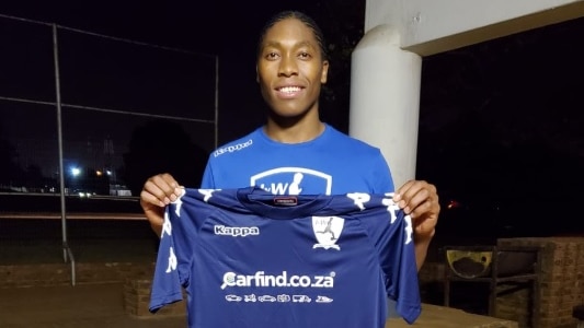 Caster Semenya stands holding a purple football shirt in front of her wearing football kit
