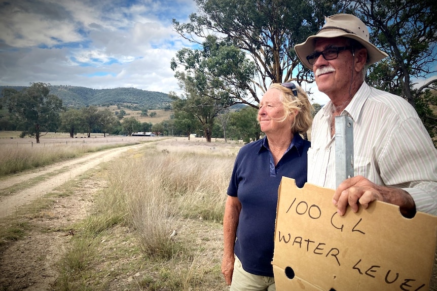 Ruth and Rob Caldwell on their driveway holding a sign that marks how far a 100 gigalitre dam would flood their property