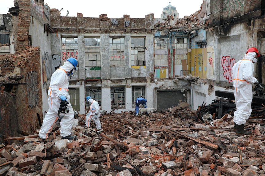 people wearing white hazmat suits walk on the debris after a fire