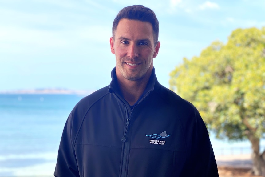 Head and shoulders photo of smiling man in navy pullover with ocean, island and tree in background, sunny day