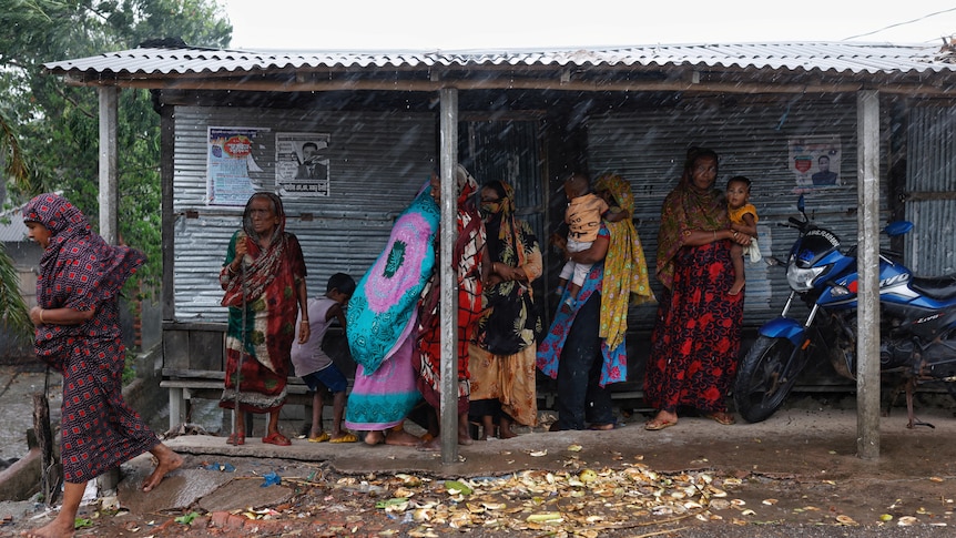 Women holding babies shelter under a tin roof as it rains.