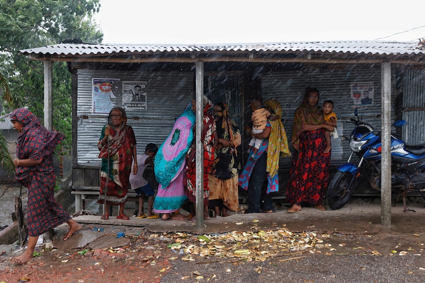 Women holding babies shelter under a tin roof as it rains.
