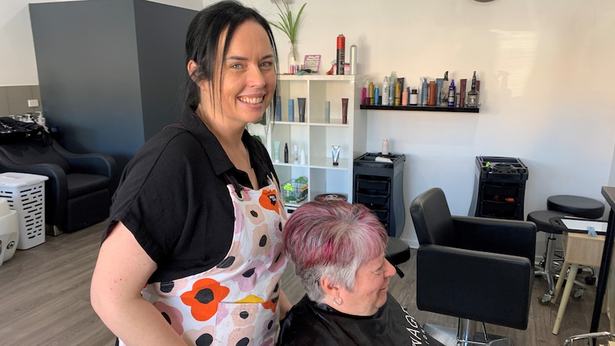 A dark haired woman wearing a floral apron standing behind a woman with pink hair seated in a beauty salon.