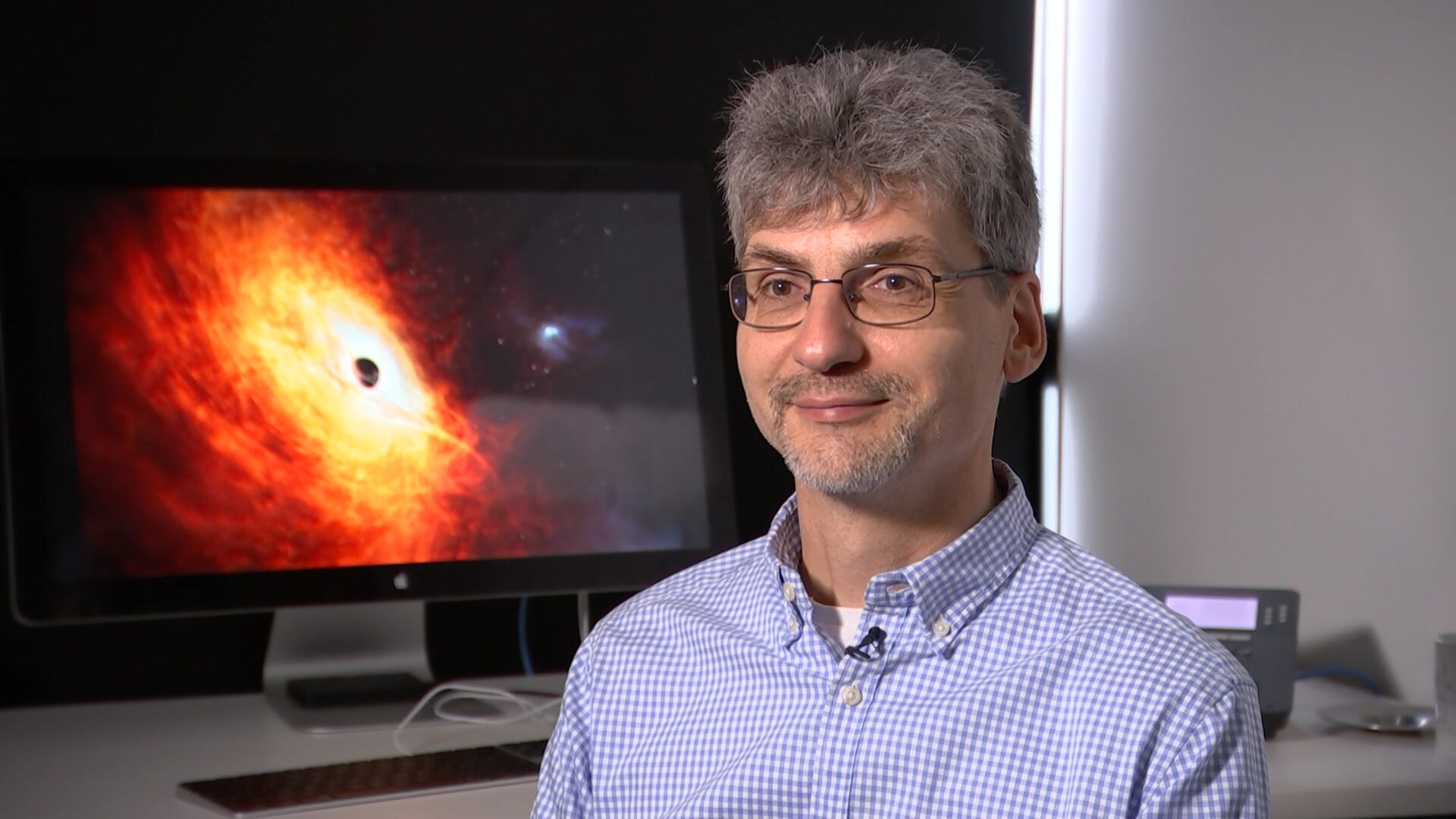 A man with grey hair and glasses smiles in front of an artist rendering of a supermassive black hole.