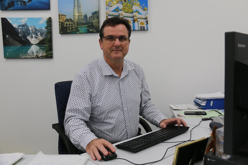 Simon sits at his desk smiling, photos of foreign destinations on the wall behind him.