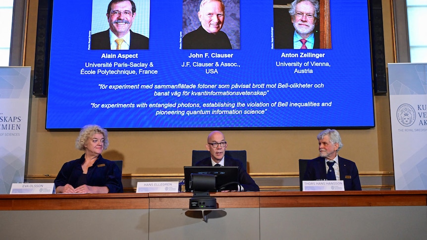 Three middle-aged people sit at a conference table in front of a screen displaying the winners of the Nobel Prize in Physics.